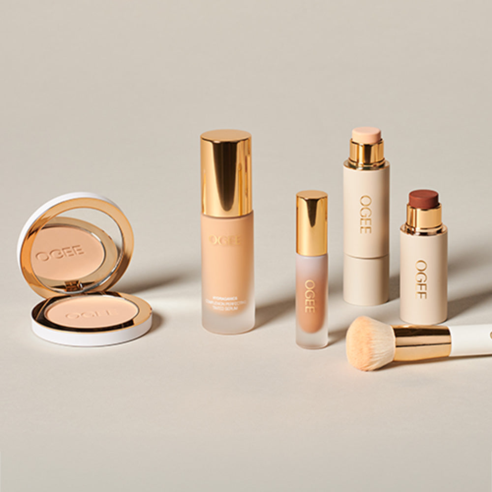 Ogee complexion products arranged on a beige surface, including an open pressed powder compact, a foundation bottle, a concealer, two face sticks, and a white-handled makeup brush.